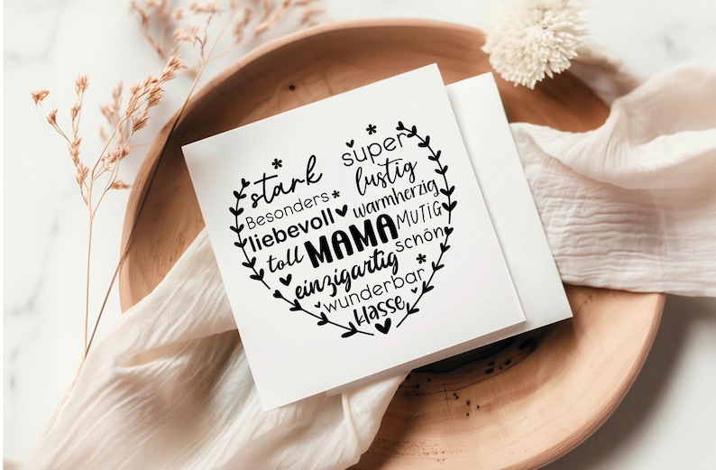 May include: A white card with a black heart-shaped design featuring the word "Mama" surrounded by positive adjectives in German. The card is on a wooden surface with a white fabric and a white flower.