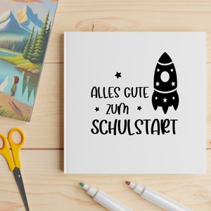 May include: A white notebook with the German text "ALLES GUTE ZUM SCHULSTART" and a rocket ship illustration. The notebook is surrounded by colorful markers, scissors, and a picture book with a landscape scene.