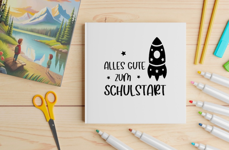 May include: A white notebook with the German text "ALLES GUTE ZUM SCHULSTART" and a rocket ship illustration. The notebook is surrounded by colorful markers, scissors, and a picture book with a landscape scene.