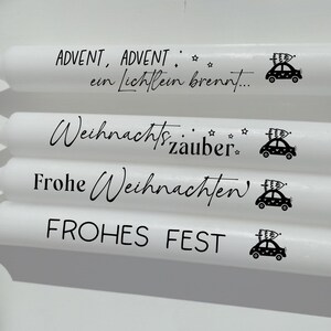 May include: Four white candles with black text and a small car graphic. The text on the candles reads "Advent, Advent, ein Lichtlein brennt...", "Weihnachtszauber", "Frohe Weihnachten", and "FROHES FEST".