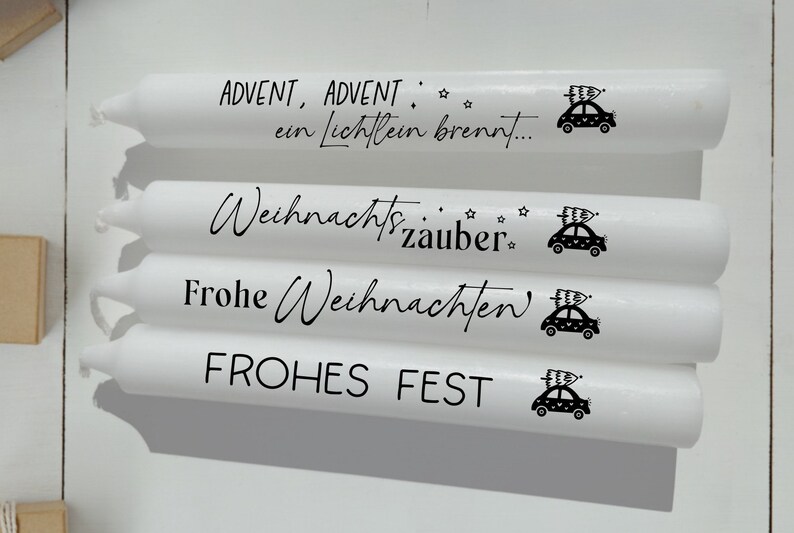 May include: Four white candles with black text and a small car graphic. The text on the candles reads "Advent, Advent, ein Lichtlein brennt...", "Weihnachtszauber", "Frohe Weihnachten", and "FROHES FEST".