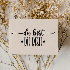 May include: A white card with black text that reads "du bist die beste" with two small black hearts on either side of the text. The card is sitting on a bed of dried white flowers.