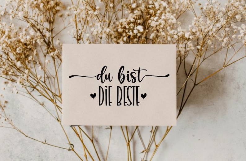 May include: A white card with black text that reads "du bist die beste" with two small black hearts on either side of the text. The card is sitting on a bed of dried white flowers.