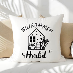 May include: White throw pillow with black text that says "Willkommen Herbst" and a black and white illustration of a house with a pumpkin and fall foliage.