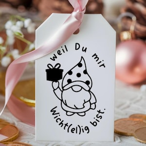 K&ouml;nnte beinhalten: Ein wei&szlig;er Geschenkanh&auml;nger mit einer schwarz-wei&szlig;en Illustration eines Zwergs, der eine Geschenkbox h&auml;lt. Der Text auf dem Anh&auml;nger lautet "weil Du mir wichtig(e)lig bist."