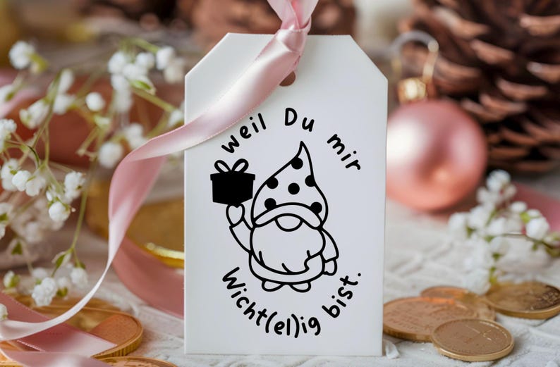 K&ouml;nnte beinhalten: Ein wei&szlig;er Geschenkanh&auml;nger mit einer schwarz-wei&szlig;en Illustration eines Zwergs, der eine Geschenkbox h&auml;lt. Der Text auf dem Anh&auml;nger lautet "weil Du mir wichtig(e)lig bist."