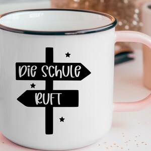 May include: White enamel mug with a black rim and a pink handle. The mug features a black signpost design with the words "Die Schule Ruft" in white, and small star accents.
