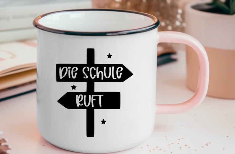 May include: White enamel mug with a black rim and a pink handle. The mug features a black signpost design with the words "Die Schule Ruft" in white, and small star accents.