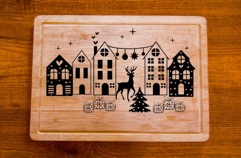 Puede incluir: Una tabla de cortar de madera con un dise&ntilde;o de pueblo navide&ntilde;o. El dise&ntilde;o presenta cinco casas, un reno, un &aacute;rbol de Navidad y regalos. Las casas son blancas y negras con detalles de coraz&oacute;n y estrella. El reno es negro y el &aacute;rbol de Navidad es verde.