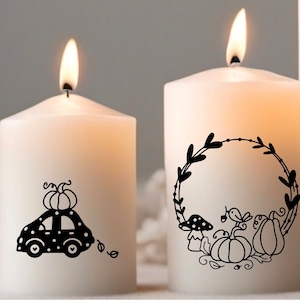 Op de afbeelding: Twee witte kaarsen met zwarte herfstthema-ontwerpen. E&eacute;n kaars heeft een auto met een pompoen erop en de andere kaars heeft een krans met pompoenen, paddenstoelen en bladeren.