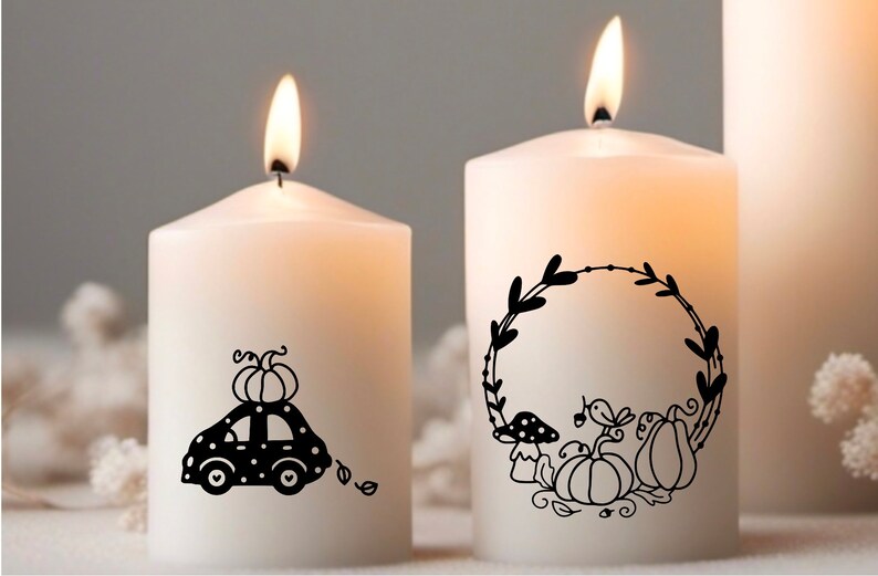 Op de afbeelding: Twee witte kaarsen met zwarte herfstthema-ontwerpen. E&eacute;n kaars heeft een auto met een pompoen erop en de andere kaars heeft een krans met pompoenen, paddenstoelen en bladeren.