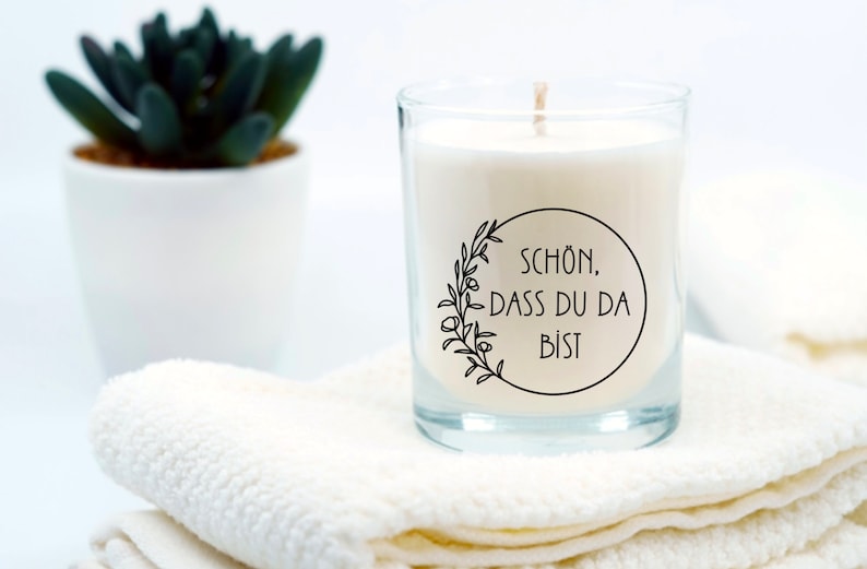 Op de afbeelding: Een witte kaars in een glazen pot met een zwart-wit label waarop "SCH&Ouml;N, DASS DU DA BIST" staat. De kaars staat op een witte handdoek.