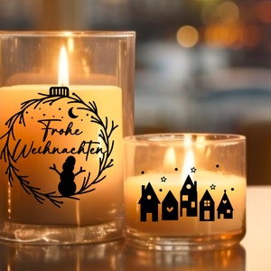 May include: Two glass candle jars with a warm glow. The larger jar has a black and white design of a wreath with the text "Frohe Weihnachten" and a snowman. The smaller jar has a black and white design of a row of houses with stars.