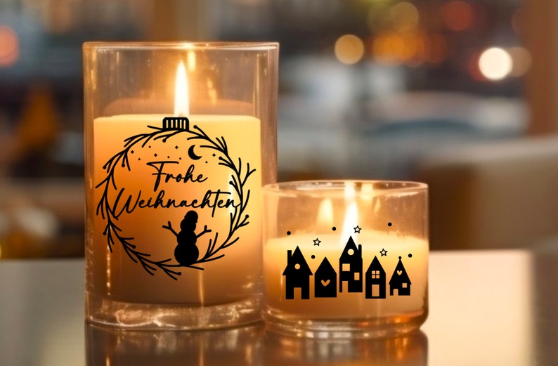 May include: Two glass candle jars with a warm glow. The larger jar has a black and white design of a wreath with the text "Frohe Weihnachten" and a snowman. The smaller jar has a black and white design of a row of houses with stars.