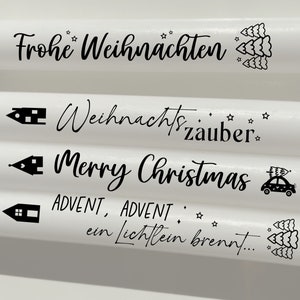 May include: Four white candles with black text and illustrations. The candles feature the phrases "Frohe Weihnachten", "Weihnachtszauber", "Merry Christmas", and "ADVENT, ADVENT, ein Lichflein brennt..."