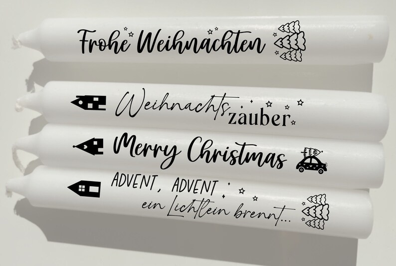 May include: Four white candles with black text and illustrations. The candles feature the phrases "Frohe Weihnachten", "Weihnachtszauber", "Merry Christmas", and "ADVENT, ADVENT, ein Lichflein brennt..."