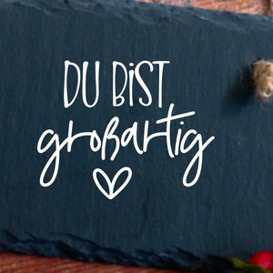May include: A black slate sign with white text that reads "DU BIST gro&szlig;artig" with a small heart underneath.