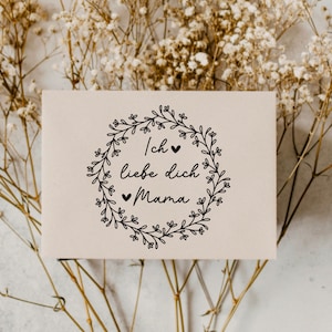 May include: A white card with a black floral wreath and the text "Ich liebe dich Mama" with small hearts.