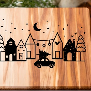 May include: A wooden cutting board with a black silhouette design of a town with houses, trees, a car carrying a Christmas tree, and a crescent moon with stars.