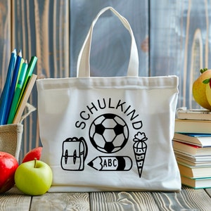 May include: White canvas tote bag with black text and illustrations. The text says "Schulkind" and the illustrations include a soccer ball, a backpack, a pencil, and a cone.