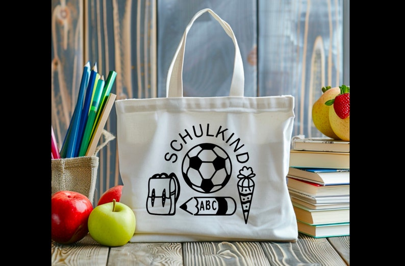 May include: White canvas tote bag with black text and illustrations. The text says "Schulkind" and the illustrations include a soccer ball, a backpack, a pencil, and a cone.