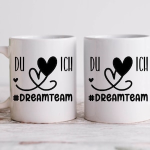 May include: Two white ceramic mugs with black handles. Each mug has a black heart design with the words "DU" and "ICH" on either side of the heart. The text "#DREAMTEAM" is printed below the heart design.