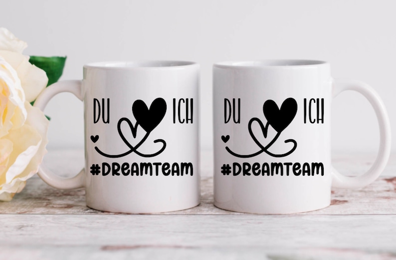 May include: Two white ceramic mugs with black handles. Each mug has a black heart design with the words "DU" and "ICH" on either side of the heart. The text "#DREAMTEAM" is printed below the heart design.