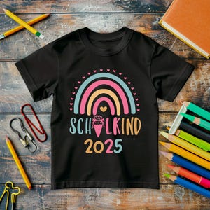 May include: Black t-shirt with a colorful rainbow design and the text "SCHULKIND 2025". The shirt is surrounded by school supplies, including pencils, paper clips, and a notebook. The rainbow features pink, orange, and blue colors.