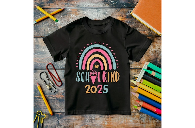 May include: Black t-shirt with a colorful rainbow design and the text "SCHULKIND 2025". The shirt is surrounded by school supplies, including pencils, paper clips, and a notebook. The rainbow features pink, orange, and blue colors.