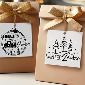 Puede incluir: Dos bolsas de regalo marrones con etiquetas de regalo blancas. La etiqueta de la bolsa de la izquierda tiene una ilustraci&oacute;n en blanco y negro de un cami&oacute;n con un &aacute;rbol de Navidad en la parte superior y el texto "Weihnachtszauber". La etiqueta de la bolsa de la derecha tiene una ilustraci&oacute;n en blanco y negro de tres &aacute;rboles y el texto "Winterzauber".