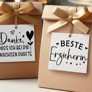 May include: Two beige gift bags with gold ribbon bows. Each bag has a white tag with black script. One says "Danke, DASS ICH BEI DIR WACHSEN DURFT" and the other says "BESTE Erzieherin".