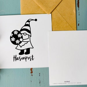 May include: A white card with a black and white illustration of a gnome holding a round object with a snowflake design. The text "Hasenpost" is written below the gnome.