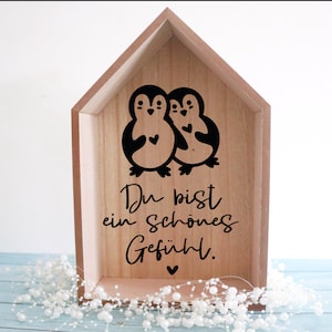 Puede incluir: Una caja de madera con forma de casa con dos ping&uuml;inos negros que sostienen corazones. El texto "Du bist ein sch&ouml;nes Gef&uuml;hl." est&aacute; escrito debajo de los ping&uuml;inos.