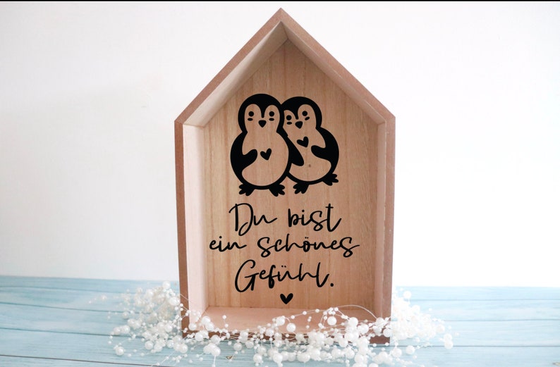 Puede incluir: Una caja de madera con forma de casa con dos ping&uuml;inos negros que sostienen corazones. El texto "Du bist ein sch&ouml;nes Gef&uuml;hl." est&aacute; escrito debajo de los ping&uuml;inos.