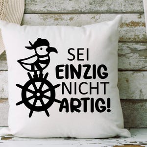 May include: White square cushion featuring a black illustration of a bird in a hat on a ship's wheel, with the German phrase "SEI EINZIG NICHT ARTIG!"