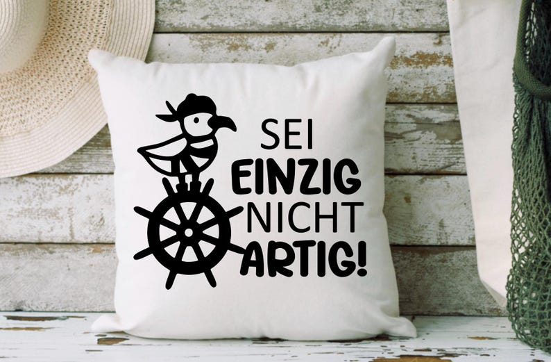 May include: White square cushion featuring a black illustration of a bird in a hat on a ship's wheel, with the German phrase "SEI EINZIG NICHT ARTIG!"