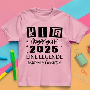 May include: A light pink t-shirt with black text that reads "KITA Abg&auml;ngerin 2025 EINE LEGENDE geht vom Gel&auml;nde." The shirt has a crew neck and short sleeves. The text is in a bold, sans-serif font.
