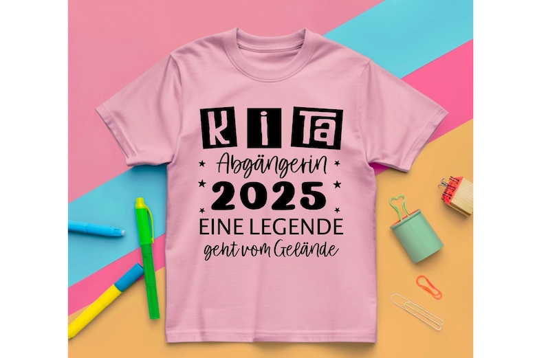 May include: A light pink t-shirt with black text that reads "KITA Abg&auml;ngerin 2025 EINE LEGENDE geht vom Gel&auml;nde." The shirt has a crew neck and short sleeves. The text is in a bold, sans-serif font.