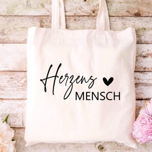 May include: White canvas tote bag with black text that reads "Herzens Mensch" with a small black heart symbol.