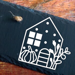 Op de afbeelding: Witte omtrek van een huis met een raam, een pompoen, een hek en een tak met bladeren op een zwarte leisteen achtergrond.