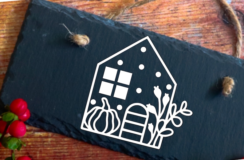 Op de afbeelding: Witte omtrek van een huis met een raam, een pompoen, een hek en een tak met bladeren op een zwarte leisteen achtergrond.