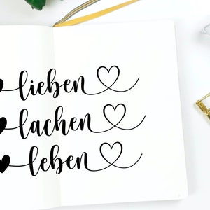 May include: A white notebook with black ink calligraphy that reads "lieben, lachen, leben" with a heart symbol before each word.