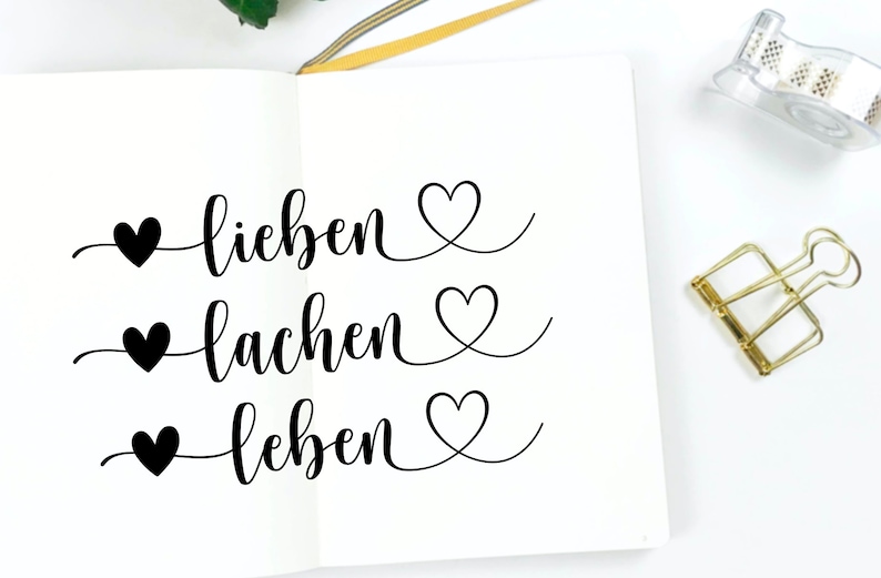 May include: A white notebook with black ink calligraphy that reads "lieben, lachen, leben" with a heart symbol before each word.