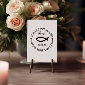 May include: A white card with a black circle design featuring a fish symbol, a cross, and the text "Sch&ouml;n dass du bist Annie 28.04.24 Meine Konfirmation". The card is on a small wooden easel.