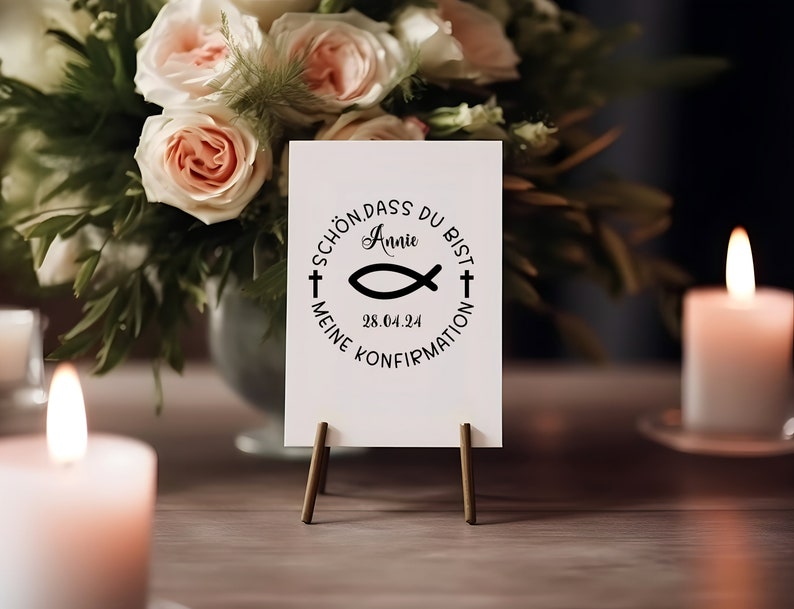 May include: A white card with a black circle design featuring a fish symbol, a cross, and the text "Sch&ouml;n dass du bist Annie 28.04.24 Meine Konfirmation". The card is on a small wooden easel.