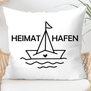 May include: White square cushion featuring the words "HEIMAT HAFEN" in black, above a line drawing of a sailboat with a heart on the hull. The cushion is on a wicker chair.