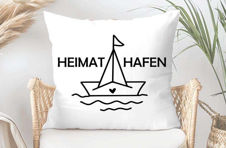 May include: White square cushion featuring the words "HEIMAT HAFEN" in black, above a line drawing of a sailboat with a heart on the hull. The cushion is on a wicker chair.
