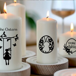 May include: Four white pillar candles with black Easter-themed designs. The candles are lit and sitting on wooden candle holders. The designs include a bunny on a swing, a bunny in a wreath, and the text "Frohe Ostern" in a wreath.
