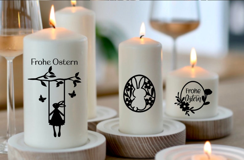 May include: Four white pillar candles with black Easter-themed designs. The candles are lit and sitting on wooden candle holders. The designs include a bunny on a swing, a bunny in a wreath, and the text "Frohe Ostern" in a wreath.