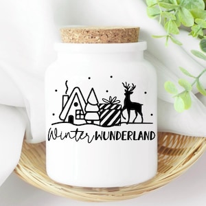 Puede incluir: Tarro de cer&aacute;mica blanco con tapa de corcho y un dise&ntilde;o de invierno de cuento de hadas en blanco y negro. El dise&ntilde;o incluye una casa, un &aacute;rbol, un regalo y un reno. El texto "Winter Wonderland" est&aacute; impreso debajo del dise&ntilde;o.